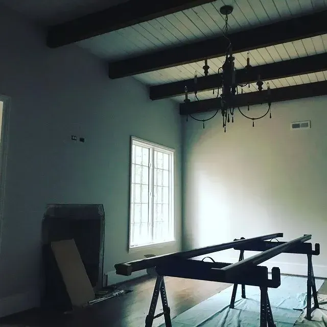 Empty room with exposed beams, chandelier, windows, and a sawhorse table in the foreground