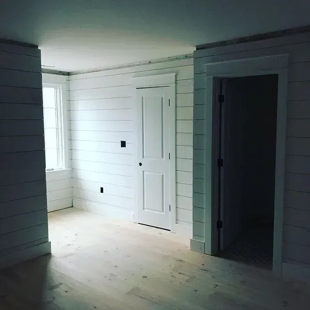 Empty white room with shiplap walls, a closed door, and a dark doorway opening on the right.