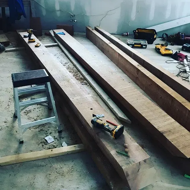 Workshop with long wooden beams, stools, and tools scattered on a concrete floor.