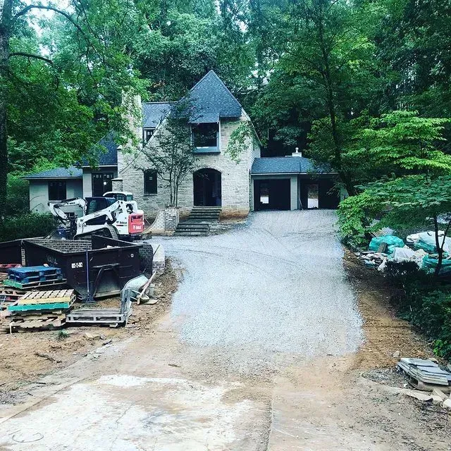 Stone house with steep roof at end of a gravel driveway, flanked by construction debris and trees