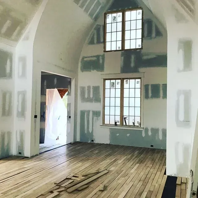 Empty unfinished room with exposed drywall, wood floorboards, and two tall windows letting in daylight