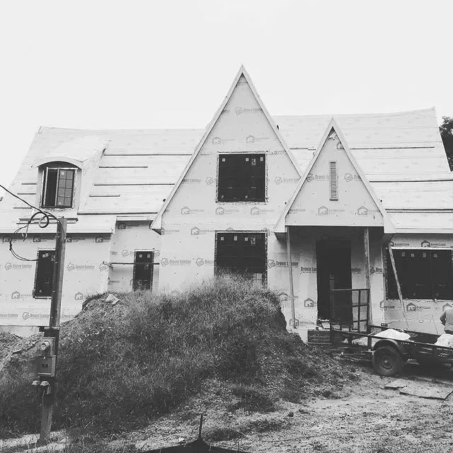 Partially built house with A-frame roofs, construction wrap, and a pile of brush in front.