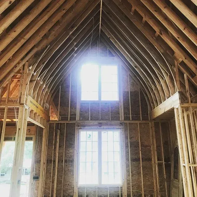Interior of a house under construction with exposed wooden rafters and bright windows at the far end