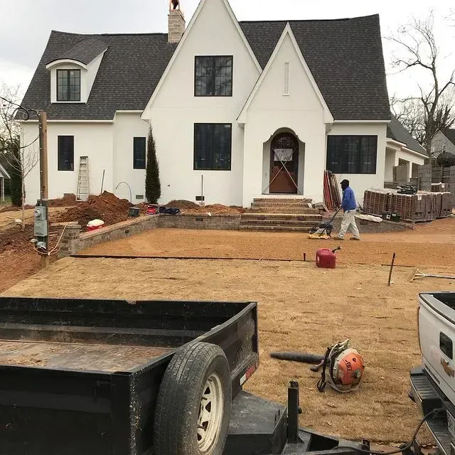 White house exterior under construction, with workers, tools, and unfinished dirt yard in front.