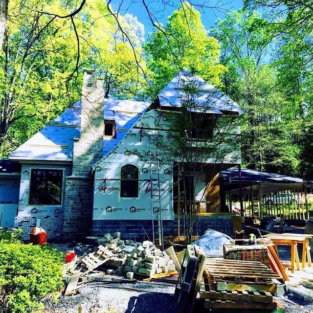Stone house under renovation surrounded by green trees, with lumber, tools, and construction materials in front