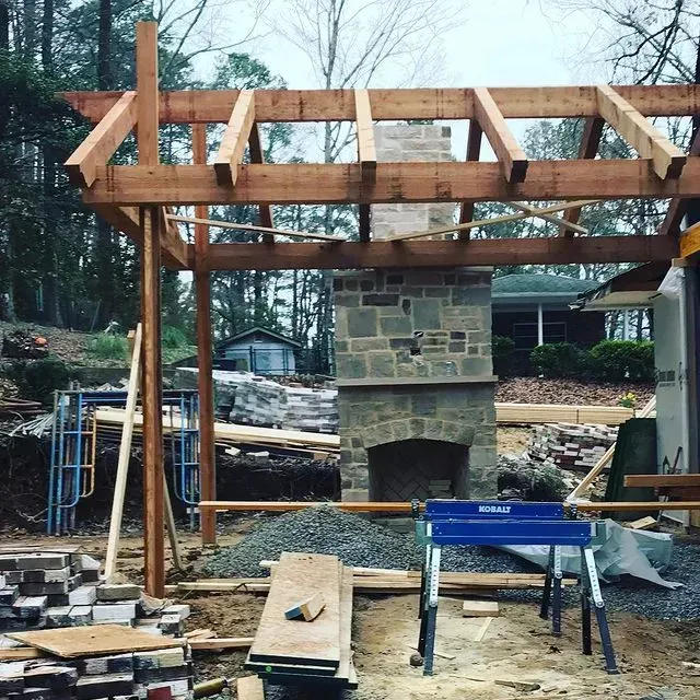 Wooden pergola under construction with a stone fireplace and blue sawhorse in a backyard.