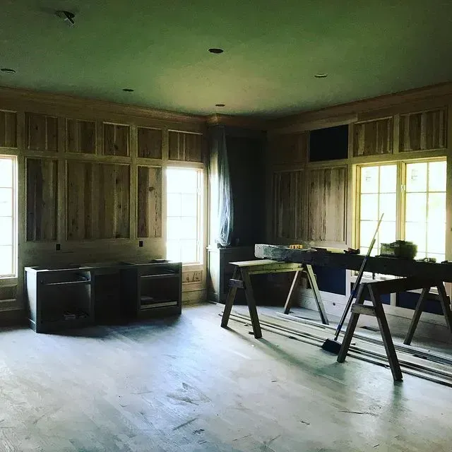 Empty rustic room with wood-paneled walls, large windows, and sawhorses holding tools near the right wall