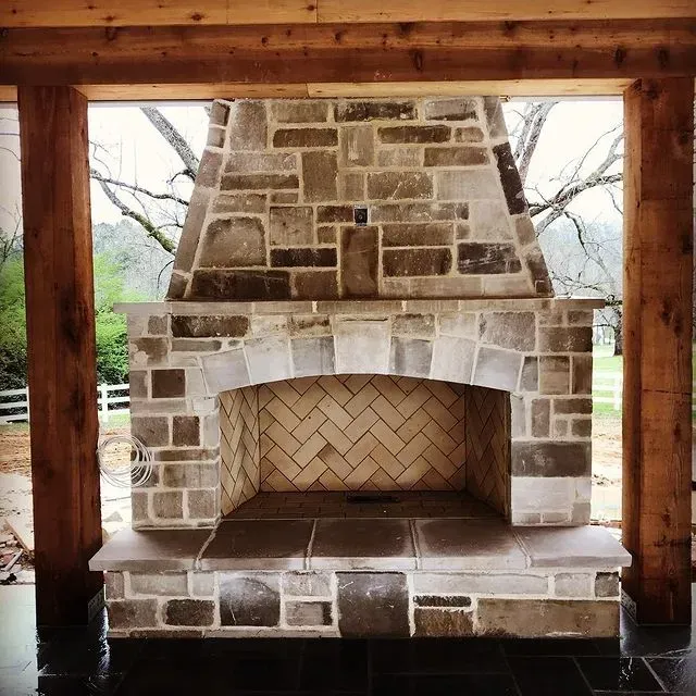 Stone outdoor fireplace with arched opening and herringbone firebrick, framed by wooden posts.