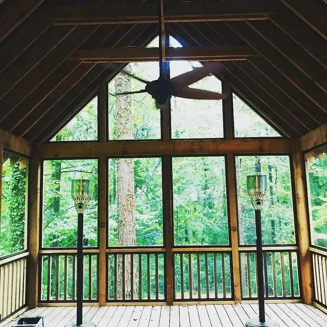Wooden screened porch with large windows, railing, ceiling fan, and lush green forest view