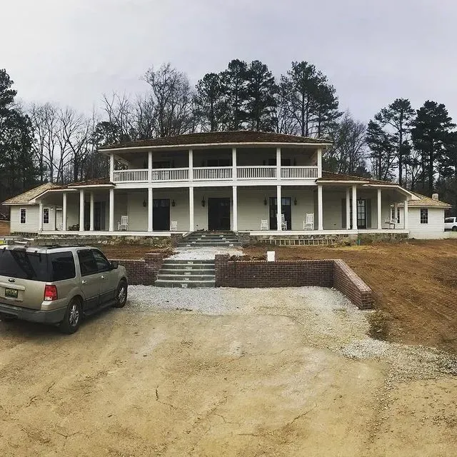 Large white two-story house with a porch, gravel driveway, and parked SUV on a wooded lot