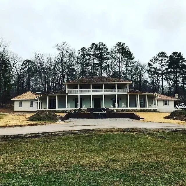 Large white house with columns and a broad porch, centered on a grassy lot with trees in the background.
