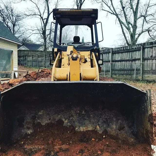Yellow front loader with a raised bucket in a fenced backyard, viewed head-on.