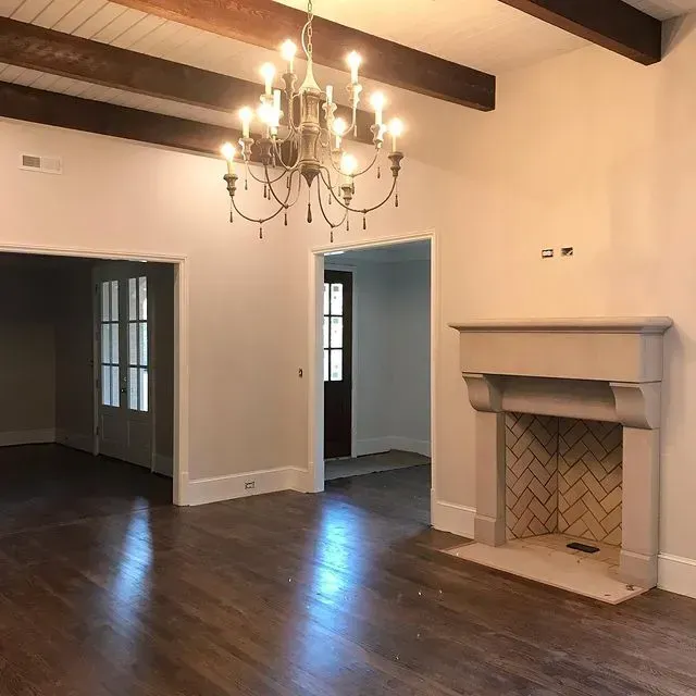 Empty living room with dark hardwood floors, exposed beams, chandelier, and fireplace mantel