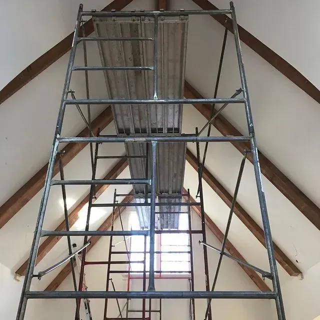 Scaffolding under a peaked roof with exposed wooden beams and hanging ductwork