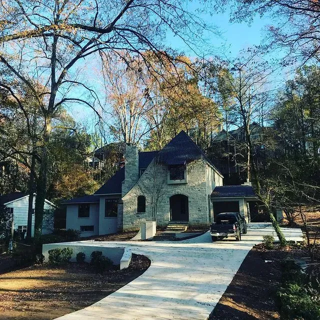 Stone house with a steep roof and driveway, surrounded by trees in autumn.