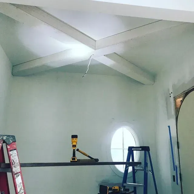 Renovation room with white walls, exposed ceiling beams, ladders, and a tool on a wall shelf.