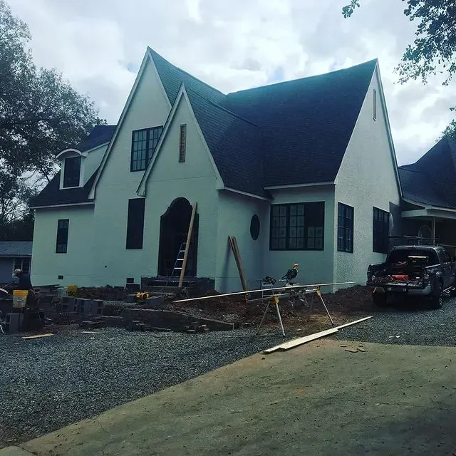 White house under construction with dark roof, gravel driveway, and a parked SUV in front
