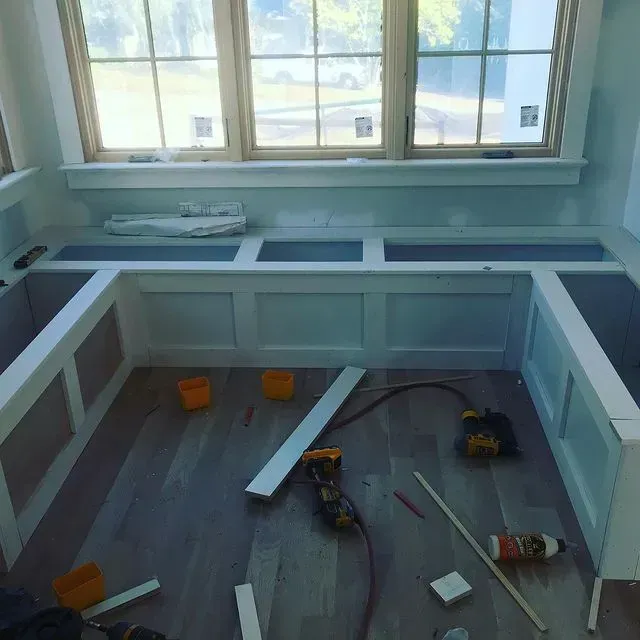 Partially built kitchen with white cabinets, gray floor, and tools scattered near a window