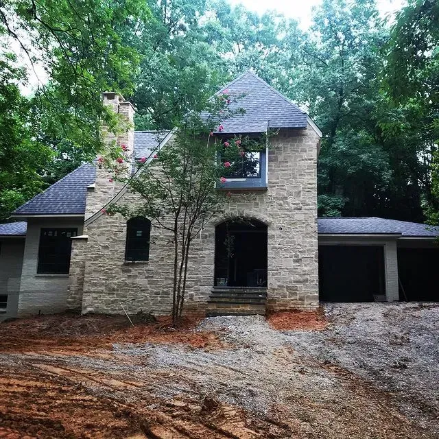 Stone house with dark roof, front porch, and garage, surrounded by trees and a gravel driveway
