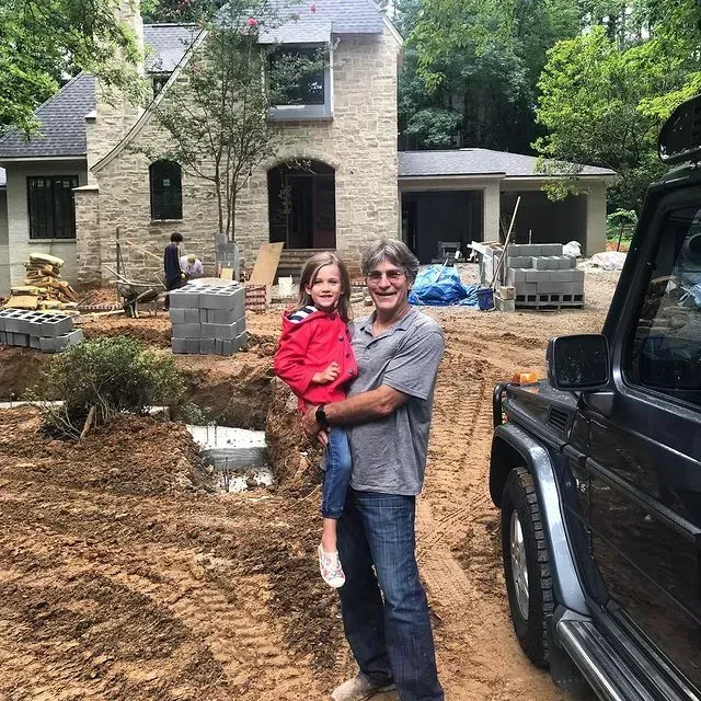 Adult holding a child in a muddy construction site in front of a stone house, with a black SUV nearby