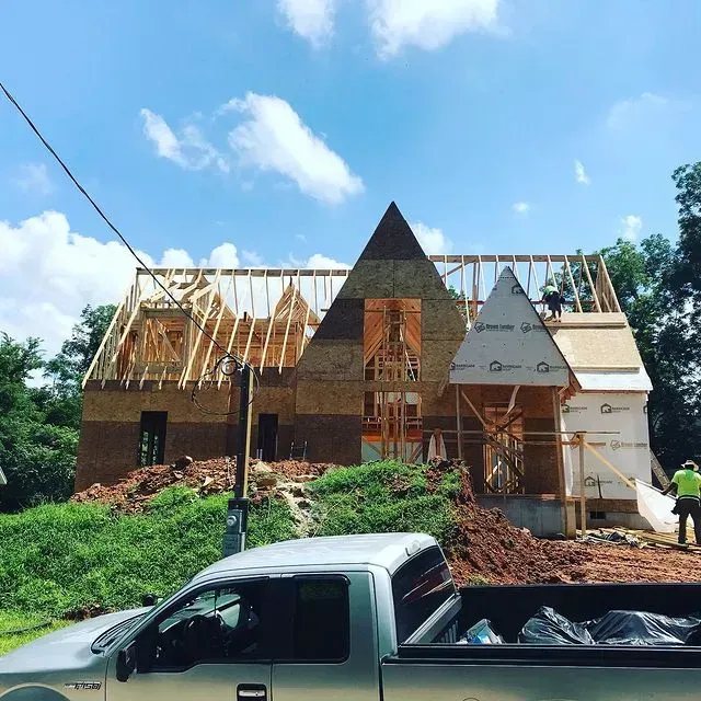 Partially built house with exposed wooden framing, a black pickup truck in front, and piles of dirt under a blue sky