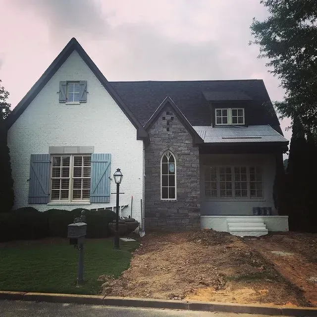 White and gray house with dark roof, front porch, small lawn, and dirt driveway under an overcast sky