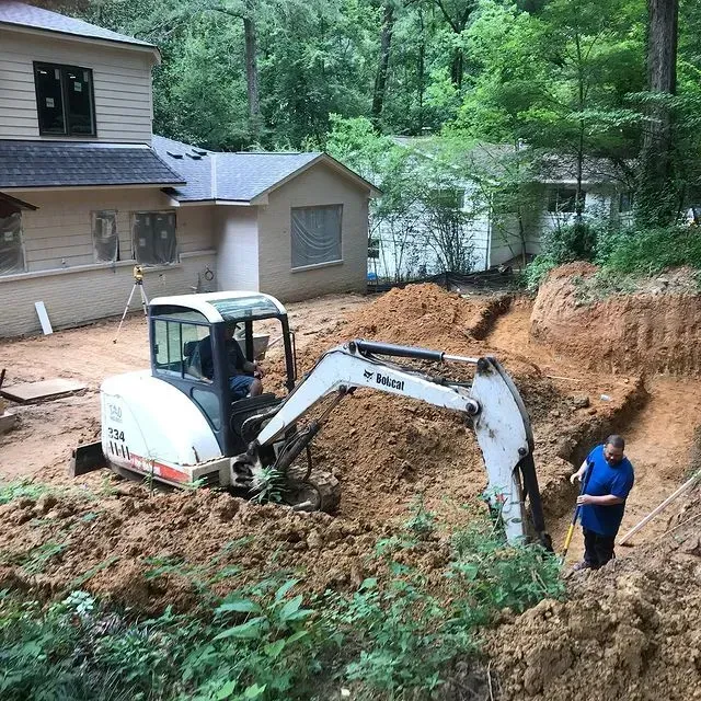 Mini excavator digging a backyard trench beside a house, with a worker standing nearby