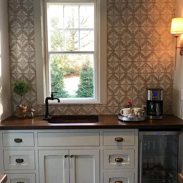 Kitchen sink and countertop with window, white cabinets, patterned wallpaper, and coffee maker.