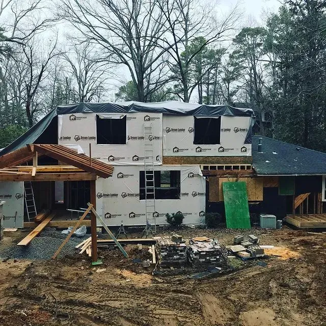 Partially built house with wrap and shingles, muddy foreground, and surrounding trees