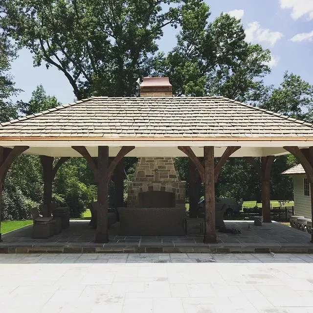 Open-air stone fireplace pavilion with wooden posts and shingled roof in a park-like setting