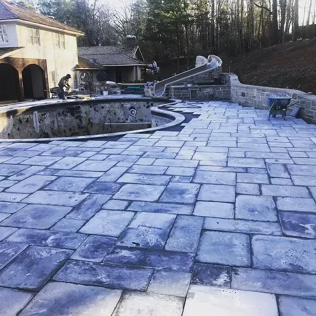 Stone patio surrounding an unfinished backyard pool, with a house and wooded hillside in the background