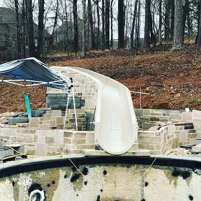 Damaged backyard slide attached to a stone retaining wall, with a collapsed canopy nearby in a wooded yard