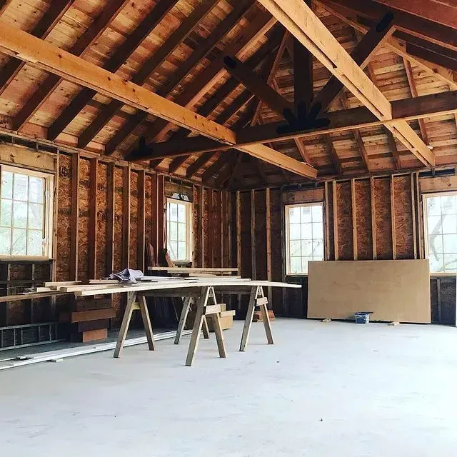 Empty unfinished room with exposed wooden framing, windows, and worktables on a white subfloor