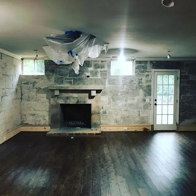 Empty room with dark wood floors, stone fireplace, and a white door; ceiling appears damaged.