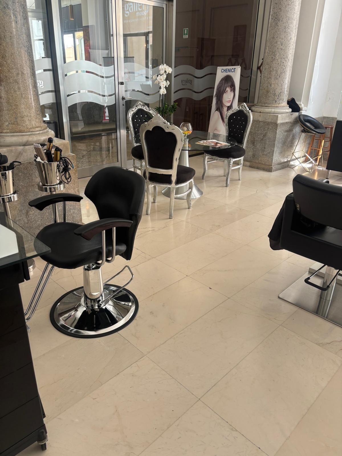 Interno di un salone con poltrone da parrucchiere nere, postazioni con specchi e pavimento in piastrelle lucide.