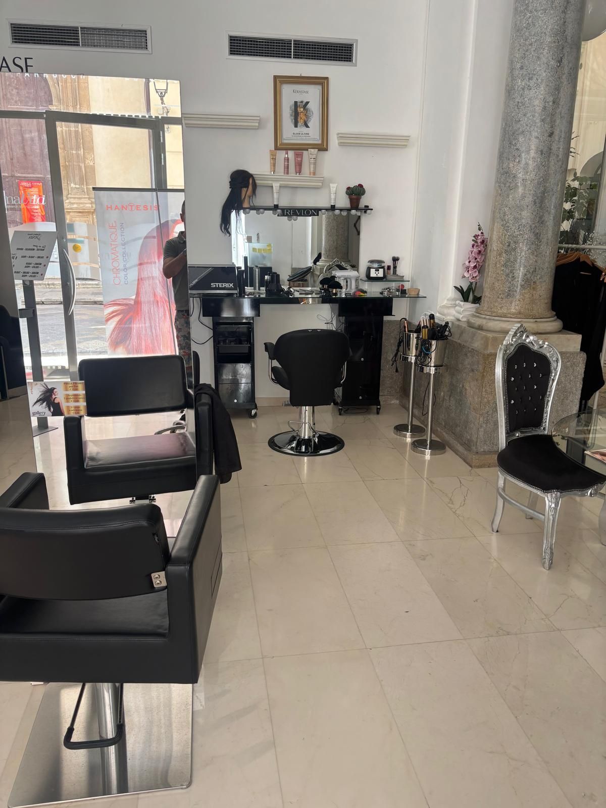 Interno di un salone da parrucchiere con sedie nere, specchi, postazioni per lo styling e pavimento piastrellato.