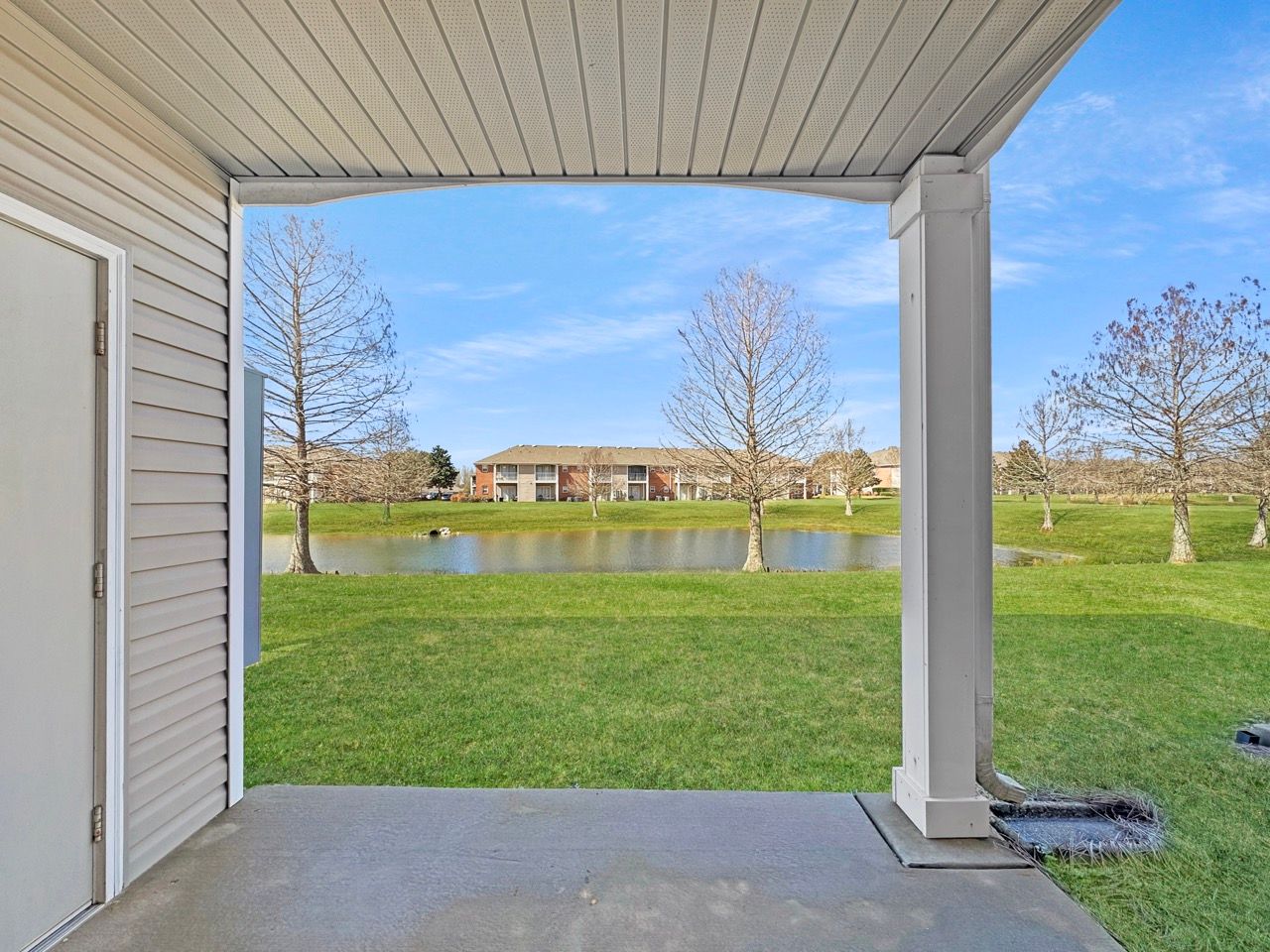 View from a covered patio: green grass, pond, apartment buildings, and a blue sky.