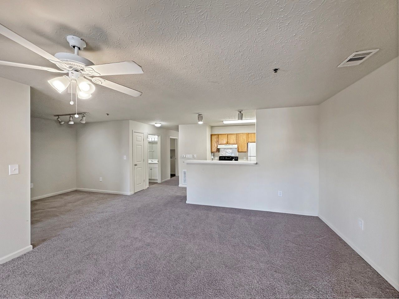 Empty living room with gray carpet, ceiling fan, and open kitchen area.