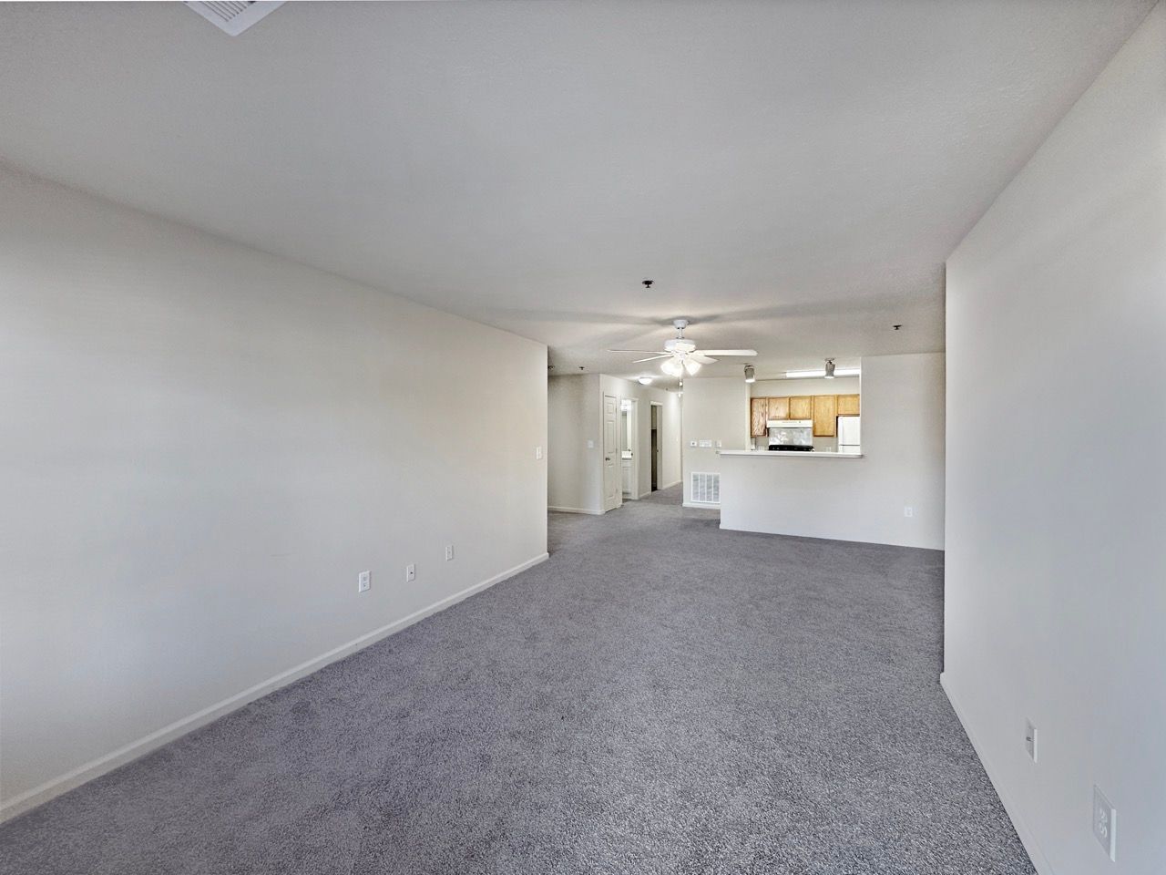 Empty living room with gray carpet, white walls, and view of the kitchen area.