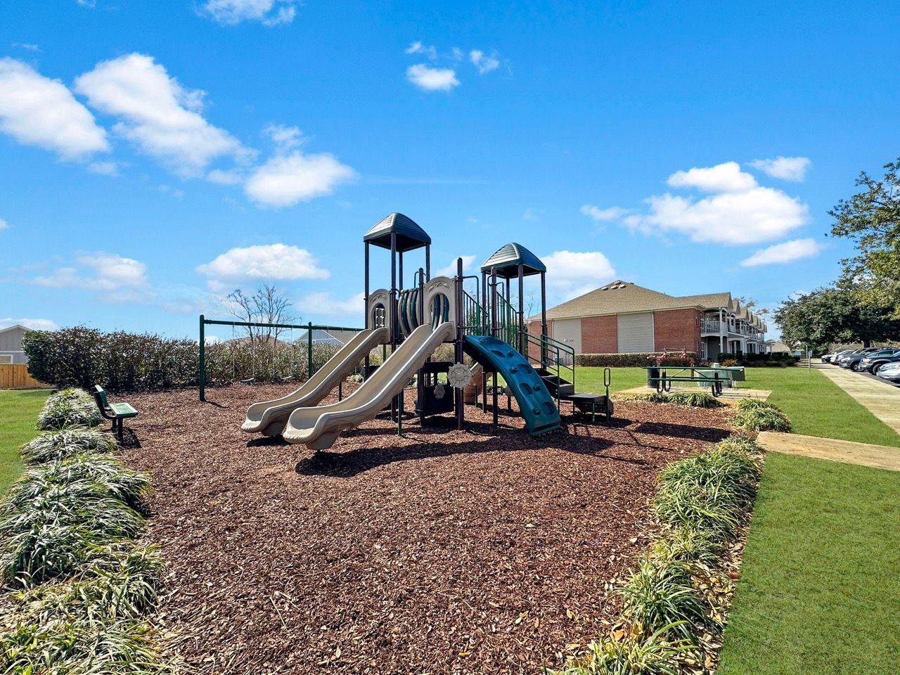 Playground with slides and structures, surrounded by mulch, green grass, and low shrubs; blue sky with clouds.