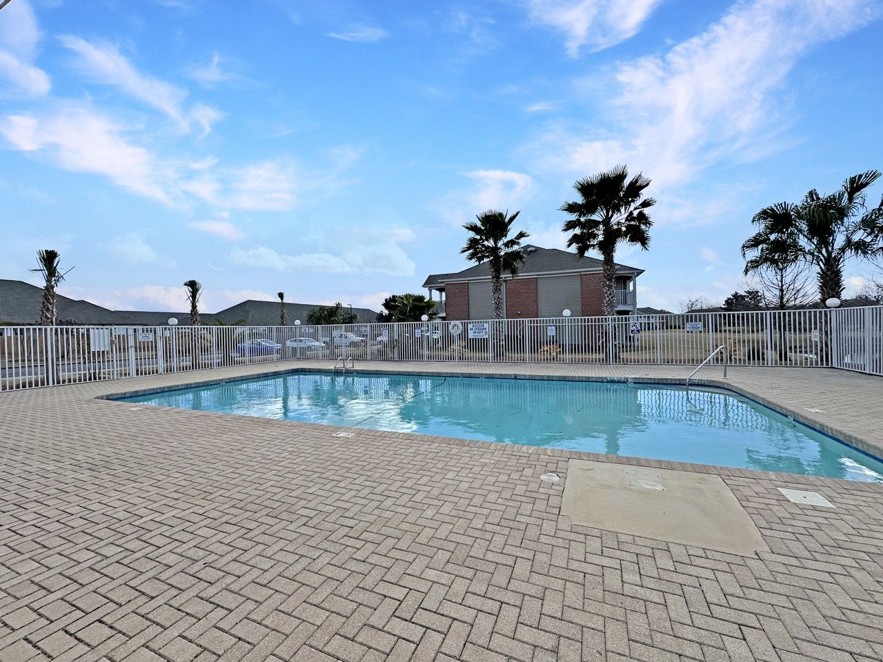 Swimming pool with brick patio, surrounded by a fence, and small building under a blue sky.