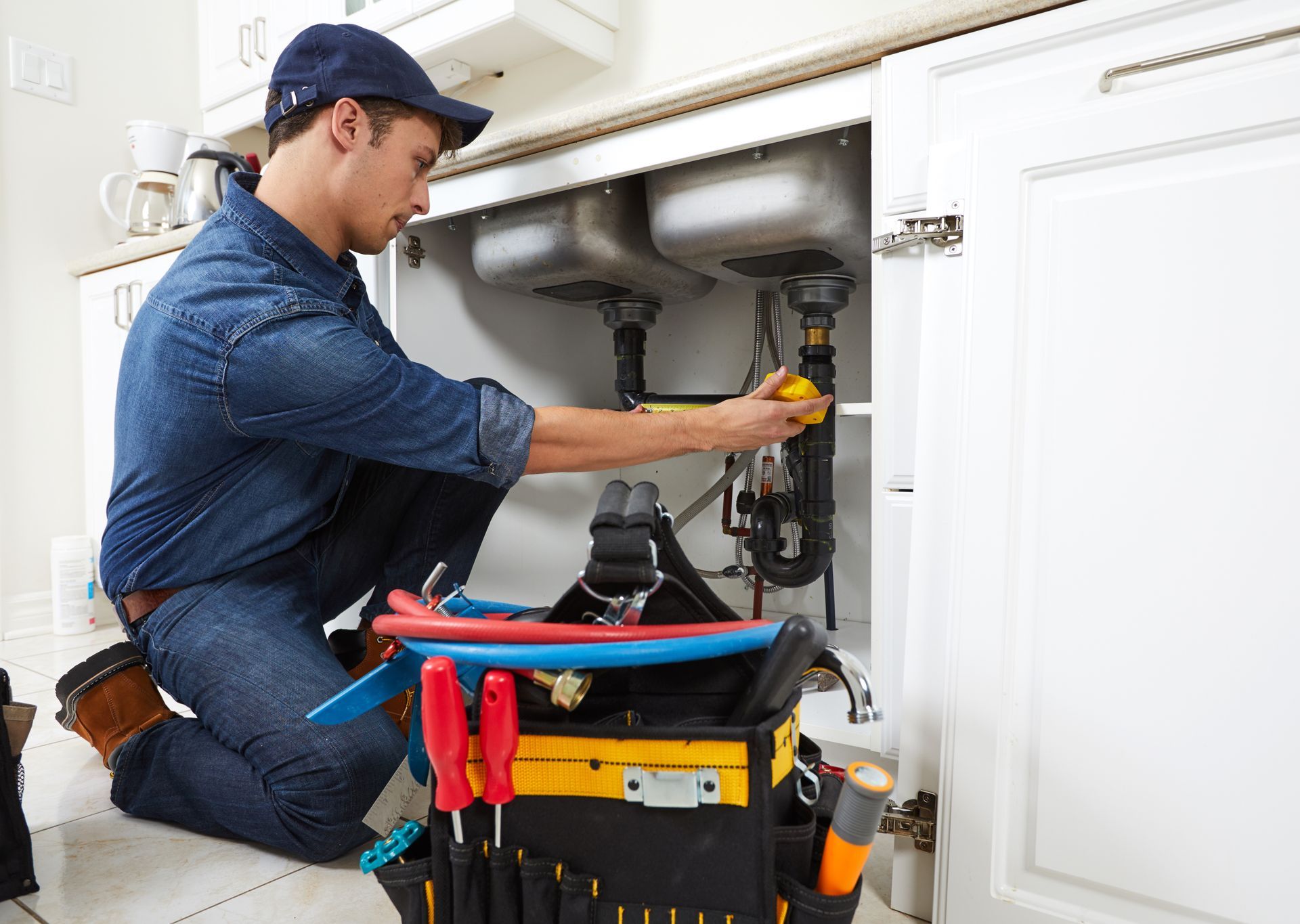 Plumber repairing pipes under a kitchen sink with tools in a toolbox nearby.