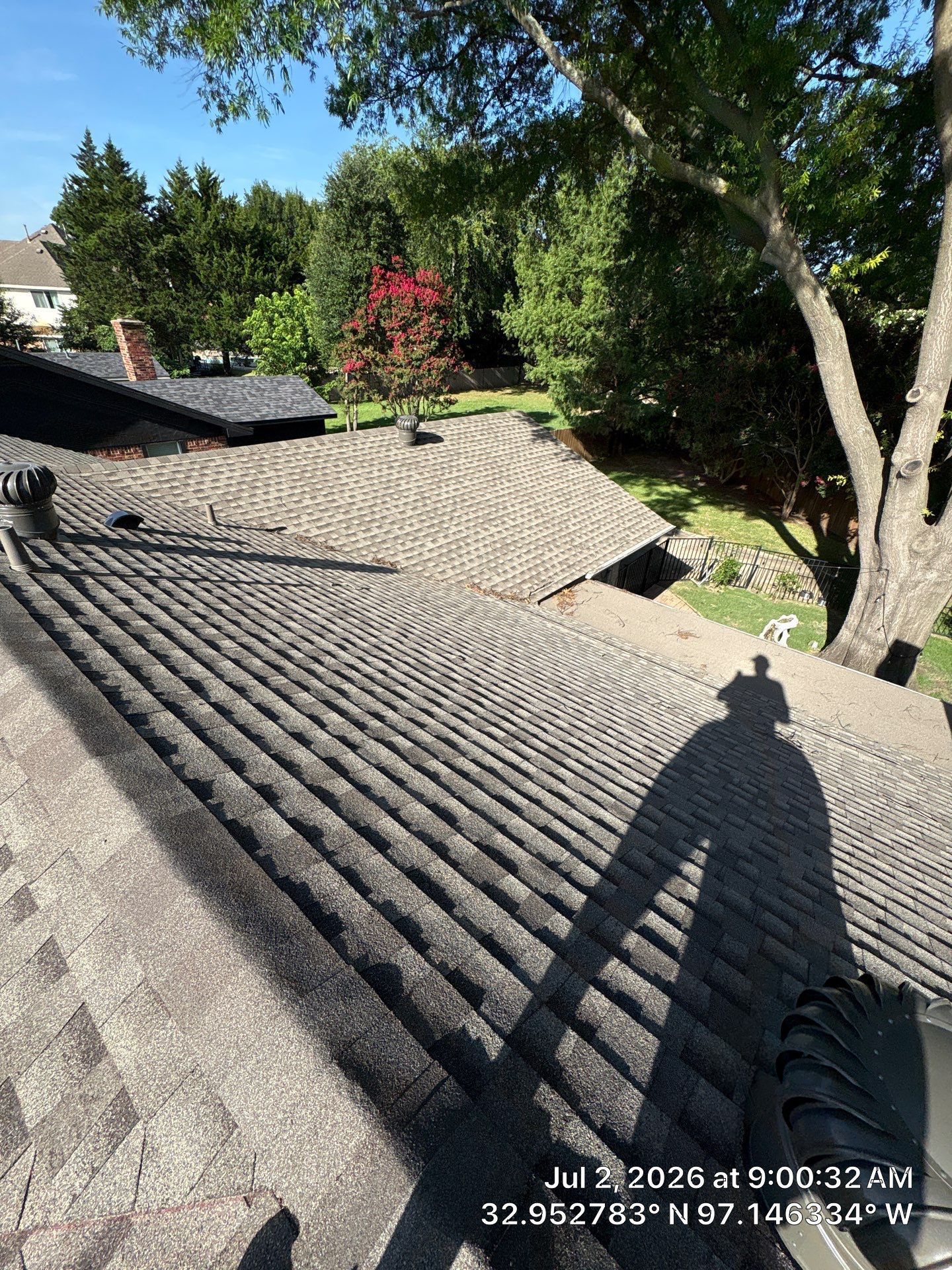 Shingled roof with trees and a bright blue sky, showing a person’s shadow on the roof.