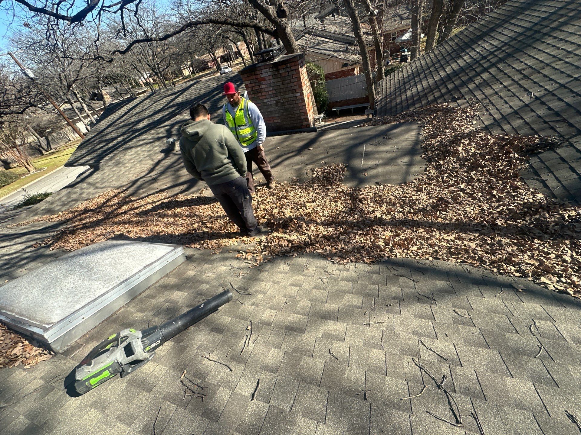Two workers in safety gear inspect a leaf-strewn rooftop beside a stone chimney and a green tool.