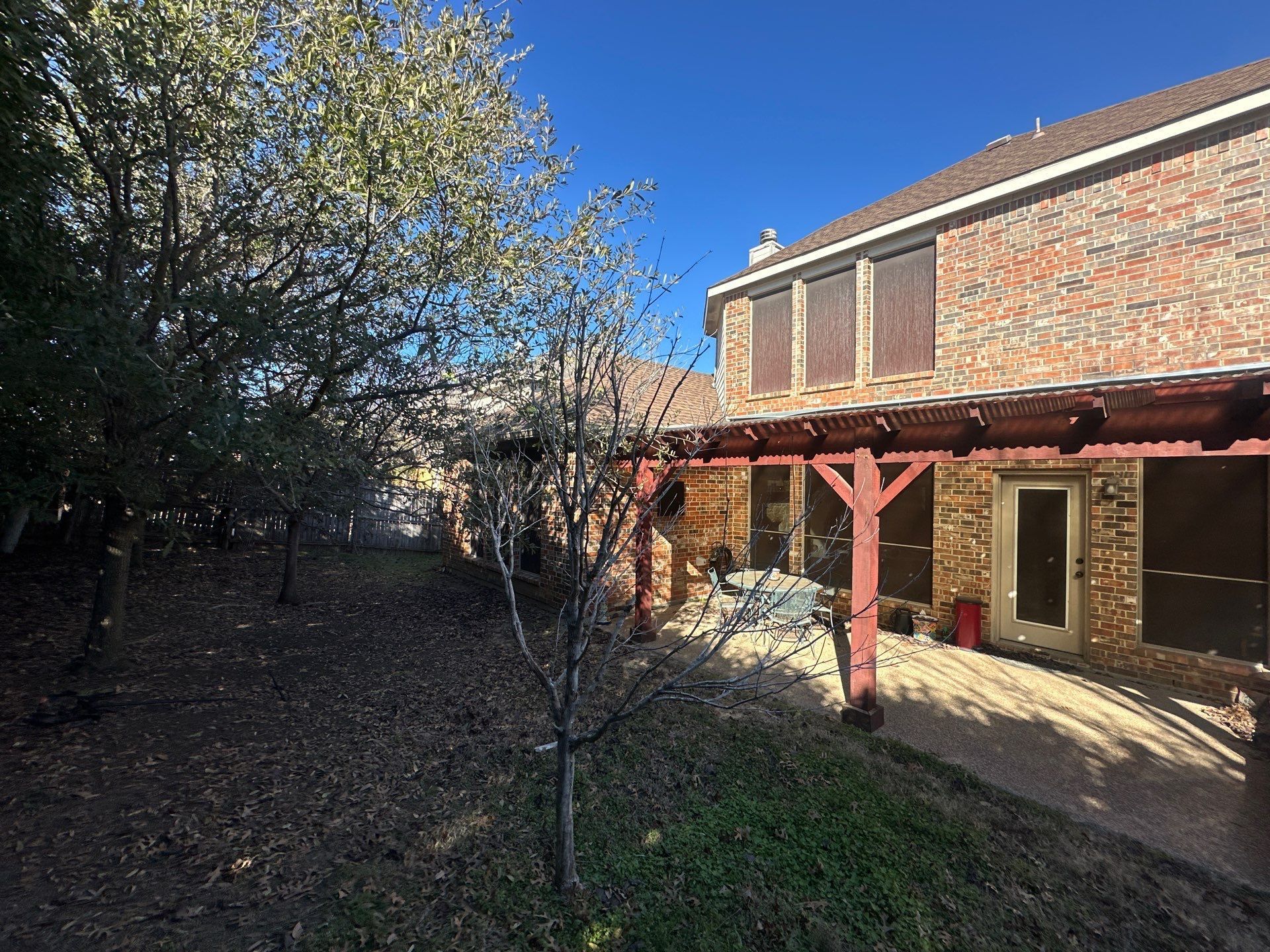 Backyard patio beside a brick house with a wooden pergola, tree, and blue sky