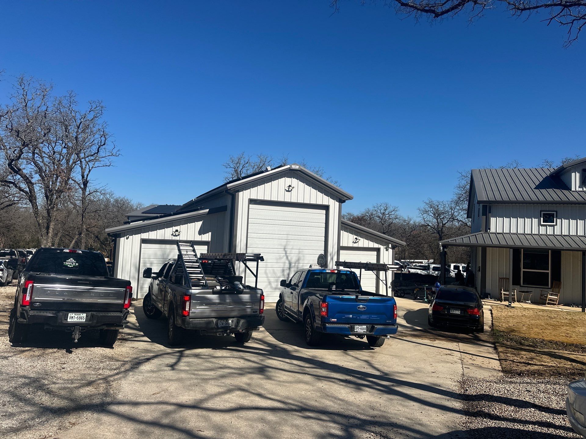 Several parked vehicles in a gravel driveway beside a white garage and house under a clear blue sky.