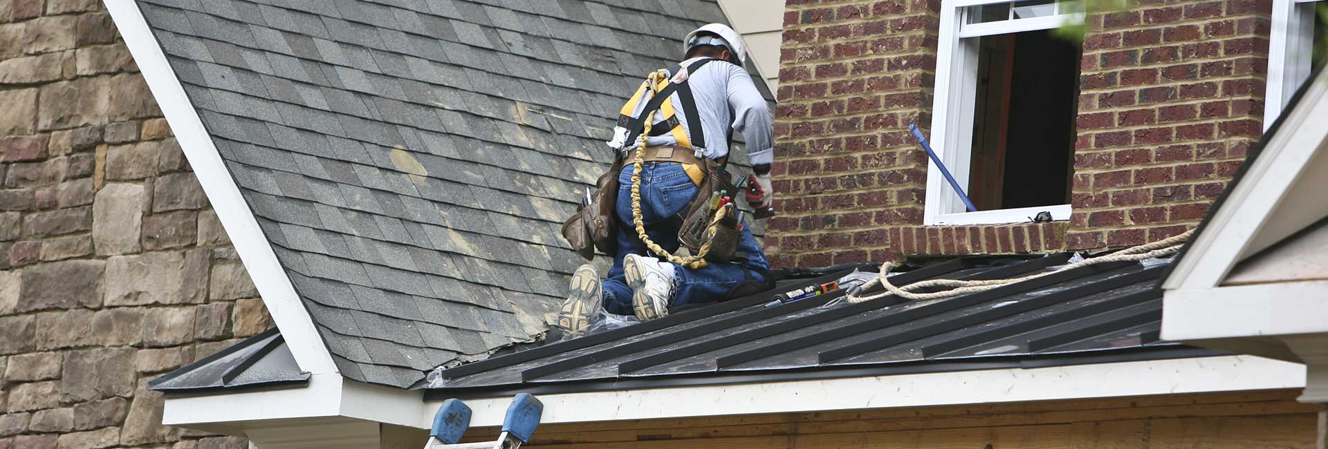 Worker in blue safety harness repairing a shingled roof beside a brick chimney and open attic window