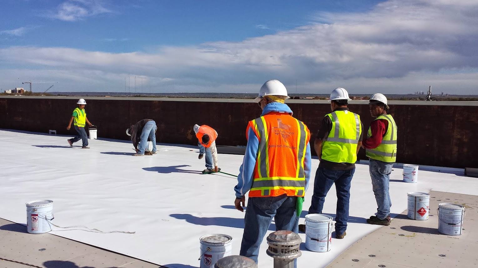 Workers in hard hats and safety vests applying white roofing material on a flat roof
