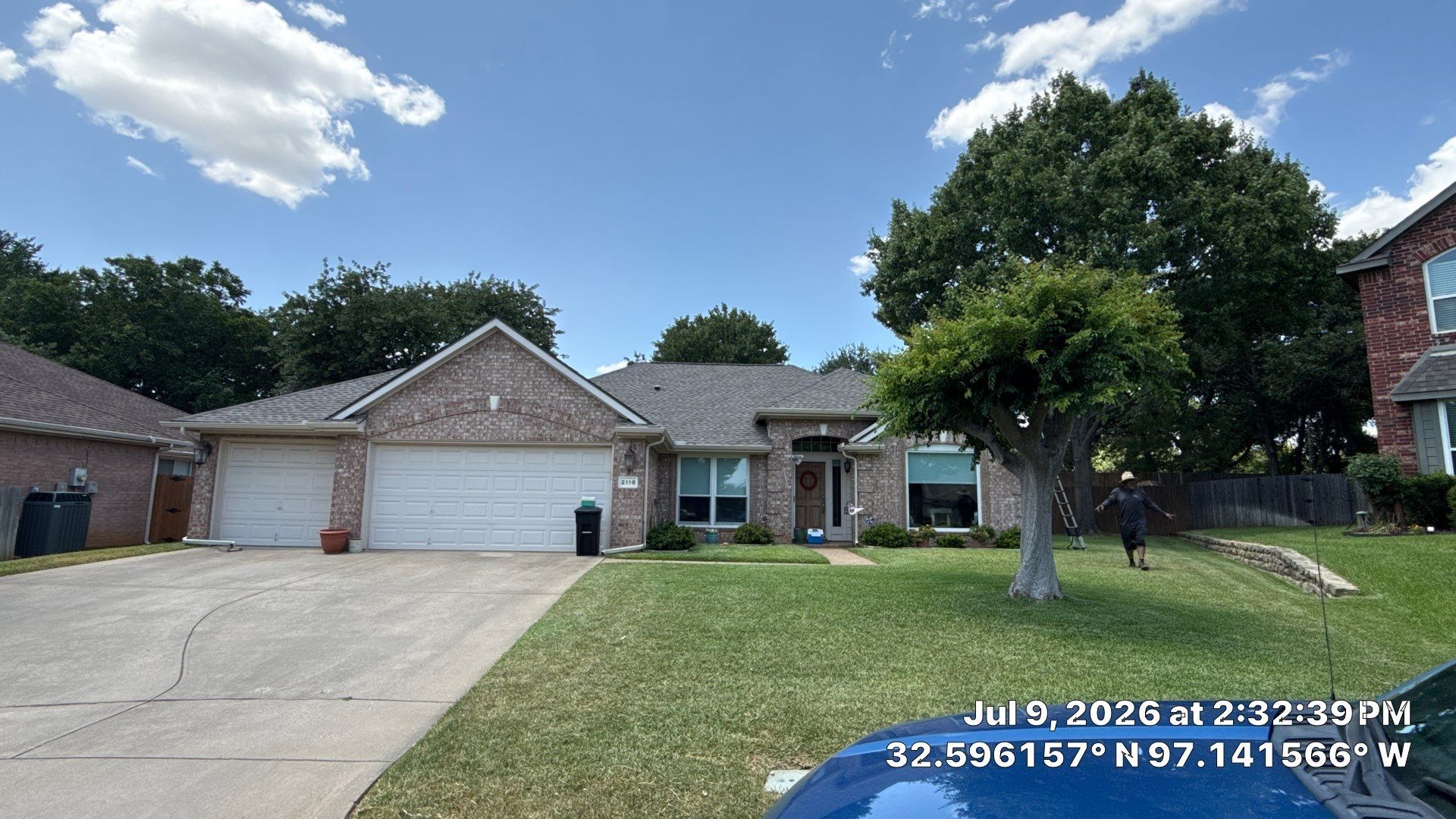 Suburban brick house with three-car garage, driveway, front lawn, and large tree under a partly cloudy sky