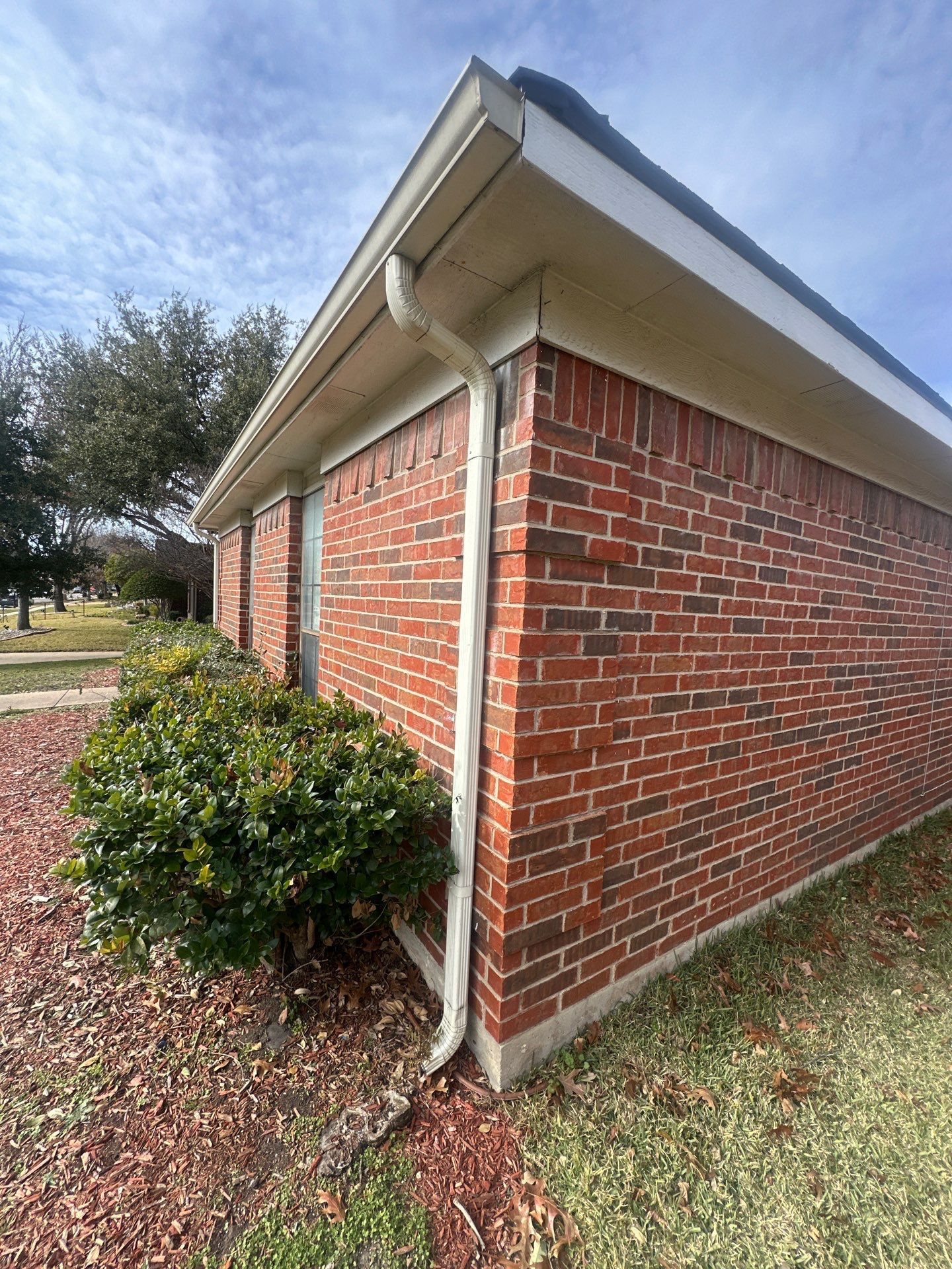 Brick building corner with white gutter, shrubs, and gravel landscaping under a cloudy sky